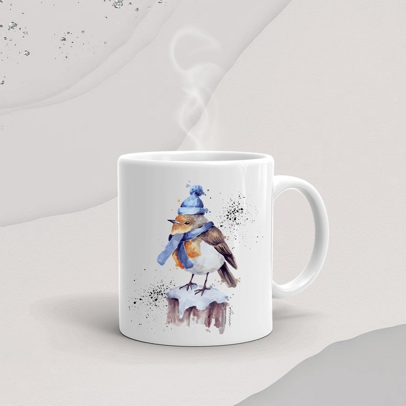 May include: White ceramic mug with a watercolor illustration of a robin wearing a blue hat and scarf, perched on a snow-covered branch. The mug has a black handle.