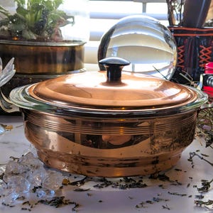 May include: A copper-colored cooking pot with a glass rim and lid, featuring a black handle on top. The pot has two metal handles on the sides. The pot is sitting on a white surface with some crystals and dried herbs.