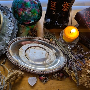 May include: An oval silver tray with a decorative rope-style rim, featuring a polished metallic surface. The tray is surrounded by dried lavender, a small yellow candle in a glass holder, a glass sphere, and a book with moon phase illustrations.