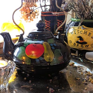 May include: Black ceramic teapot with a gold handle and a painted design of a red apple, yellow pear, and green leaves. A small candle in a glass holder is in the foreground. A cauldron with the words "Witches Brew" is in the background.