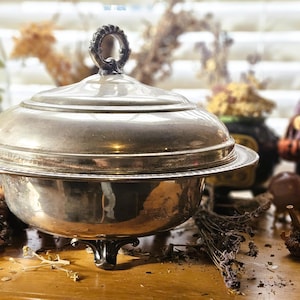 May include: A silver-toned metal serving dish with a matching domed lid and a decorative ring-shaped handle on top. The bowl features a wide, flat rim with a beaded edge and rests on three ornate, curved feet, showcasing a polished metallic finish.