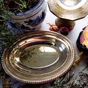 May include: A silver oval serving platter with a rope-like border. The platter reflects the surrounding objects, including a blue and white floral pot, a candle, and dried flowers. The scene has a vintage, rustic aesthetic.