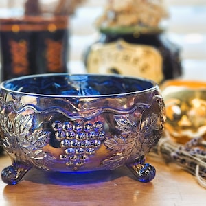 May include: A deep blue iridescent glass bowl featuring a raised relief pattern of grape clusters and vine leaves. The bowl has a scalloped rim and is supported by three small, curved feet, showcasing a reflective, metallic finish.