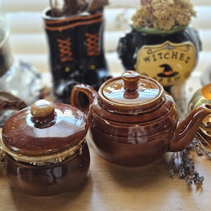 May include: A glossy brown ceramic teapot with a matching lid and a separate small brown ceramic lidded jar. Both items feature a smooth, reflective finish and are positioned together on a surface, showcasing their rounded, functional design.