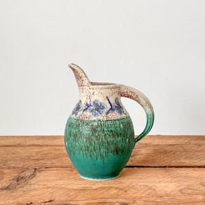 May include: A handmade ceramic pitcher with a unique design. The pitcher features a teal green base, a speckled off-white and blue upper section, and a curved handle. The spout is elongated and the pitcher sits on a wooden surface.