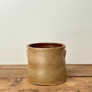 May include: A tan stoneware crock with a dark brown interior. The cylindrical antique kitchenware has two small handles. It is displayed on a wooden surface. The crock is a vintage piece.