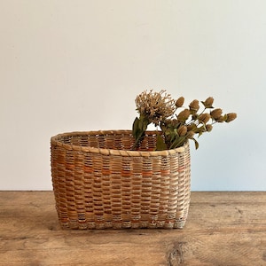 Può includere: Un cesto ovale intrecciato di colore marrone chiaro e arancione. Il cesto è fatto di materiali naturali e ha un aspetto rustico. È pieno di fiori secchi.