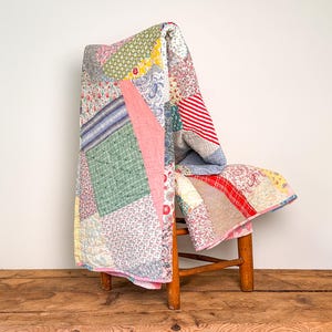 May include: A patchwork quilt featuring a variety of fabric patterns including floral prints, stripes, and geometric designs in shades of pink, green, blue, red, and yellow. The quilt is draped over a wooden chair with visible stitching.