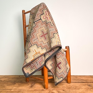 May include: A woven textile blanket featuring a geometric pattern of interlocking zig-zag lines in shades of muted red, beige, brown, and cream. The blanket has a dark grey border and is draped over a wooden chair with a natural finish.