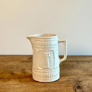 May include: A white ceramic jug with an embossed design. The jug has a handle and a spout.