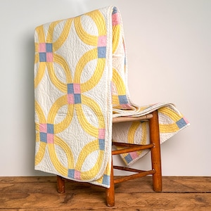 May include: A quilted blanket featuring a double wedding ring pattern in yellow, light blue, and soft pink on a white background. The fabric has a textured, stitched surface and is draped over a wooden chair with four legs and a woven seat.