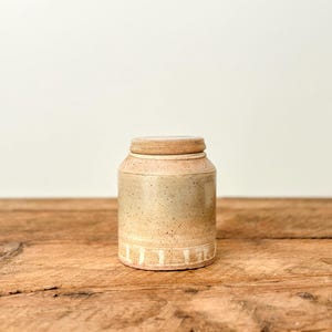 以下が含まれることがあります： お揃いの蓋が付いた、まだら模様のベージュ色の陶器の瓶。瓶は円筒形で、底の近くに白い縦線が入っています。瓶は木製の表面に置かれています。