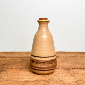 May include: A beige ceramic bottle with a narrow neck and a cork stopper. The body of the bottle features a smooth, speckled finish on the upper portion and a series of dark brown horizontal bands encircling the lower section of the vessel.