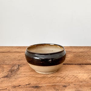 Peut inclure: Bol en céramique fait à la main avec une bande marron foncé au milieu. Le bol a une base et un intérieur beiges, et est posé sur une surface en bois. Le bol est probablement utilisé pour servir des aliments ou comme objet décoratif.