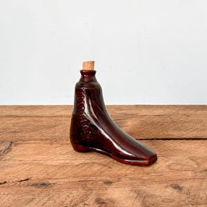 Pode incluir: Uma garrafa de cerâmica castanha em forma de bota com uma rolha de cortiça. A garrafa tem um acabamento brilhante e está colocada sobre uma superfície de madeira e um fundo branco. A garrafa em forma de bota é um item decorativo.