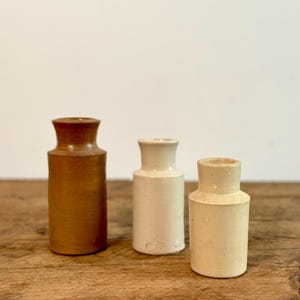 May include: Three small ceramic vases, one brown and two white, arranged on a wooden surface.