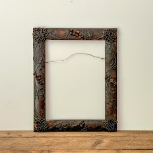 Puede incluir: Un marco de fotos rectangular y ornamentado con un acabado marrón oscuro. El marco presenta detalles florales y de vid, con racimos de bayas y flores en las esquinas y a los lados. Un alambre cruza el centro vacío.