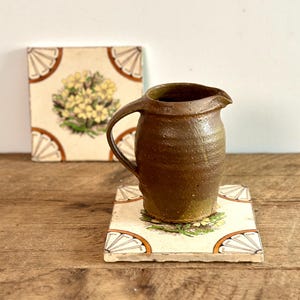 May include: A small brown ceramic jug with a handle sits on a white ceramic tile with a floral design. The tile has a brown border and a white background.