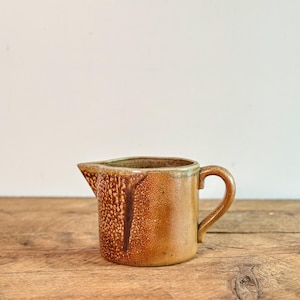 Op de afbeelding: Een kleine, bruine keramische creamer met een handvat en een tuit. De creamer heeft een textuur met een druppelachtig patroon. De creamer staat op een houten oppervlak.