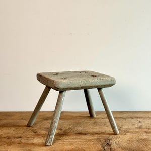 Puede incluir: Un pequeño taburete de madera rústico con tres patas. El taburete está pintado de color gris claro y tiene una parte superior cuadrada.