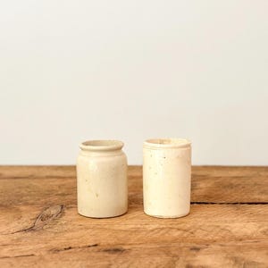 Könnte beinhalten: Zwei cremefarbene Keramikbehälter. Einer hat einen abgerundeten Körper mit einem leicht ausgestellten Rand. Der andere ist zylindrisch mit einem geraden Rand. Beide stehen auf einer Holzoberfläche.
