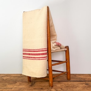 May include: A cream-colored blanket with red stripes is draped over a wooden chair. The blanket is folded over the back of the chair and hangs down the front. The chair has a woven seat and a simple, rustic design.