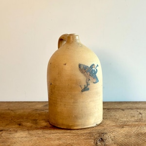 Op de afbeelding: Een vintage aardewerken kruik met een blauw en wit ontwerp van een vogel met een trompet. De kruik heeft een handvat en is lichtbruin.