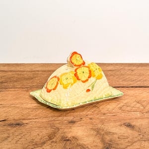 May include: A ceramic butter dish with a dome-shaped lid featuring a hand-painted floral pattern of orange and yellow flowers on a cream-coloured background. The rectangular base has a green rim, and the lid includes a small flower-shaped handle.