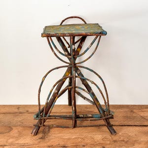 May include: A handmade, rustic stool with a small rectangular seat. The frame is made from curved, weathered metal rods and wooden pieces, painted with blue, yellow, and red accents. The stool sits on a wooden surface.