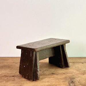 May include: A small, weathered wooden step stool with a rectangular top and four legs. The stool is painted in a dark, muted green colour, showing signs of wear and age. The stool is placed on a wooden surface.