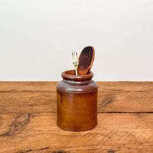May include: A cylindrical brown stoneware crock with a glossy finish, containing a small silver-toned metal fork and a dark wooden spoon. The vessel features a thick rim and a slightly tapered base, showcasing a rustic, earthy aesthetic.