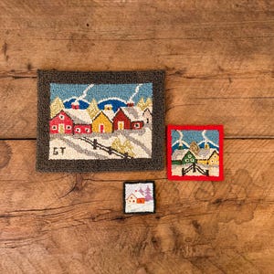 Op de afbeelding: Drie kleine, ingelijste naaldkunstwerken met winterse taferelen. De grootste toont rode en gele huizen met besneeuwde daken, een hek en de initialen "GT". De andere twee tonen vergelijkbare besneeuwde landschappen.
