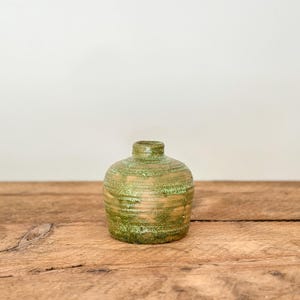 May include: A small, round ceramic vase with a narrow neck. The vase has a textured surface with a blend of green and tan colours. It sits on a rustic wooden surface against a neutral background.