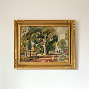 May include: A framed landscape painting featuring a tree-lined road with a building in the distance. The artwork is in a gold ornate frame. The painting depicts a sunny day with green trees and a light-colored road.
