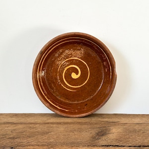 May include: A rustic, brown ceramic bowl with a spiral design in the center. The bowl has a slightly uneven rim and a glossy finish. The spiral is painted in a lighter shade of yellow. The bowl is sitting on a wooden surface.