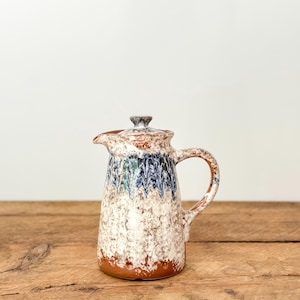 May include: A ceramic jug with a lid, featuring a handle and spout. The jug has a textured white base with blue and green accents, and a brown rim and base. The lid is a matching colour and design. The jug is on a wooden surface.