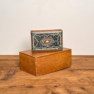Puede incluir: Una caja rectangular de madera con acabado marrón claro y textura agrietada, coronada por una caja más pequeña y ornamentada con una incrustación oscura de patrones intrincados y una piedra ovalada central con bandas concéntricas.