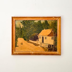 Puede incluir: Una pintura de paisaje enmarcada que representa una cabaña blanca con techo color canela situada junto a un camino de tierra. La escena incluye un denso bosque de árboles verdes, una valla de madera y una pequeña figura roja en el camino, todo en un marco de madera.