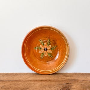May include: A round, orange ceramic plate with a floral design in the center. The flower has tan petals, green leaves, and a dark blue center. The plate has a glossy finish and is set against a white background.
