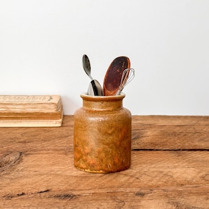May include: A rustic, brown ceramic utensil holder with a textured surface. It contains a metal spoon, a wooden spoon, and a whisk. A stack of old books sits to the left on a wooden surface. The background is a plain white wall.
