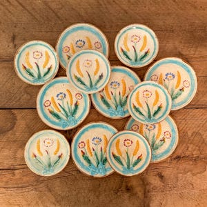 Può includere: Una collezione di undici piatti rotondi in ceramica con un disegno dipinto a mano di spighe di grano gialle e piccoli fiori blu legati con un nastro azzurro. Ogni piatto presenta un centro color crema e un bordo azzurro.