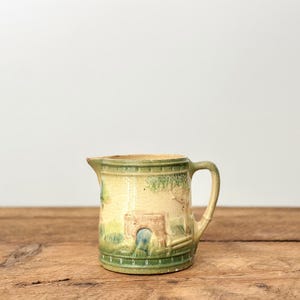 Może przedstawiać: Mały, zabytkowy ceramiczny dzbanek z uchwytem i wylewką. Dzbanek przedstawia scenę krajobrazową z mostem i drzewami, namalowaną w odcieniach zieleni, beżu i błękitu. Dzbanek stoi na drewnianej powierzchni.