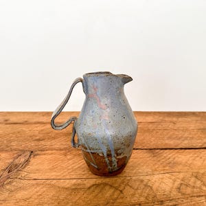 May include: A ceramic jug with a light blue, speckled glaze and a brown base. The jug features a distinct, angular handle that extends outwards and loops back to the body, with a small spout at the top rim for pouring liquids.