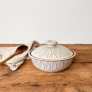 May include: A white ceramic serving bowl with a lid, featuring a decorative pattern. A wooden spoon rests on a linen cloth beside the bowl, all set on a rustic wooden surface. The bowl is approximately 20 cm in diameter.