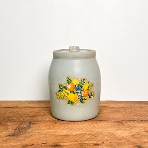 May include: A light gray ceramic crock with a matching lid, featuring a central decal of assorted fruits including yellow grapes, oranges, and dark blue berries with green leaves. The cylindrical container has a smooth, matte finish.