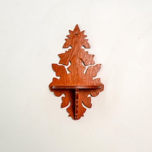 May include: A wall-mounted wooden shelf featuring a medium brown finish and a symmetrical, ornate cutout design. The back panel displays intricate, leaf-like scrollwork patterns, while a small horizontal platform is supported by a central bracket.