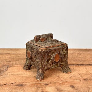 May include: A rustic, brown wooden box with a lid, designed to resemble a tree trunk. The box has a textured surface, four short legs, and a small branch detail on top. The box is sitting on a wooden surface.
