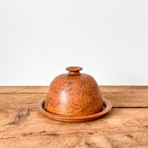 May include: A speckled, brown ceramic butter dish with a domed lid and a small knob on top. The dish sits on a matching round plate. The butter dish is made of ceramic and has a rustic, handmade appearance.