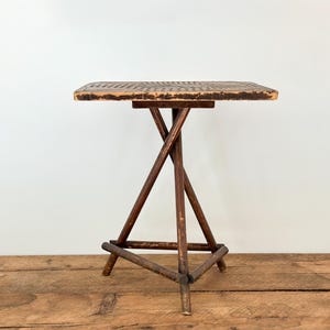 May include: A small, antique wooden side table with a square top and a unique, crossed-leg base. The tabletop has a textured surface. The table is made of dark brown wood and stands on a wooden floor.