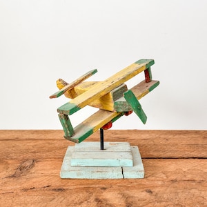 May include: A vintage wooden aeroplane sculpture with yellow, green, and red paint. The toy plane is mounted on a light blue base and stands on a wooden surface. The aeroplane has a propeller and wings.